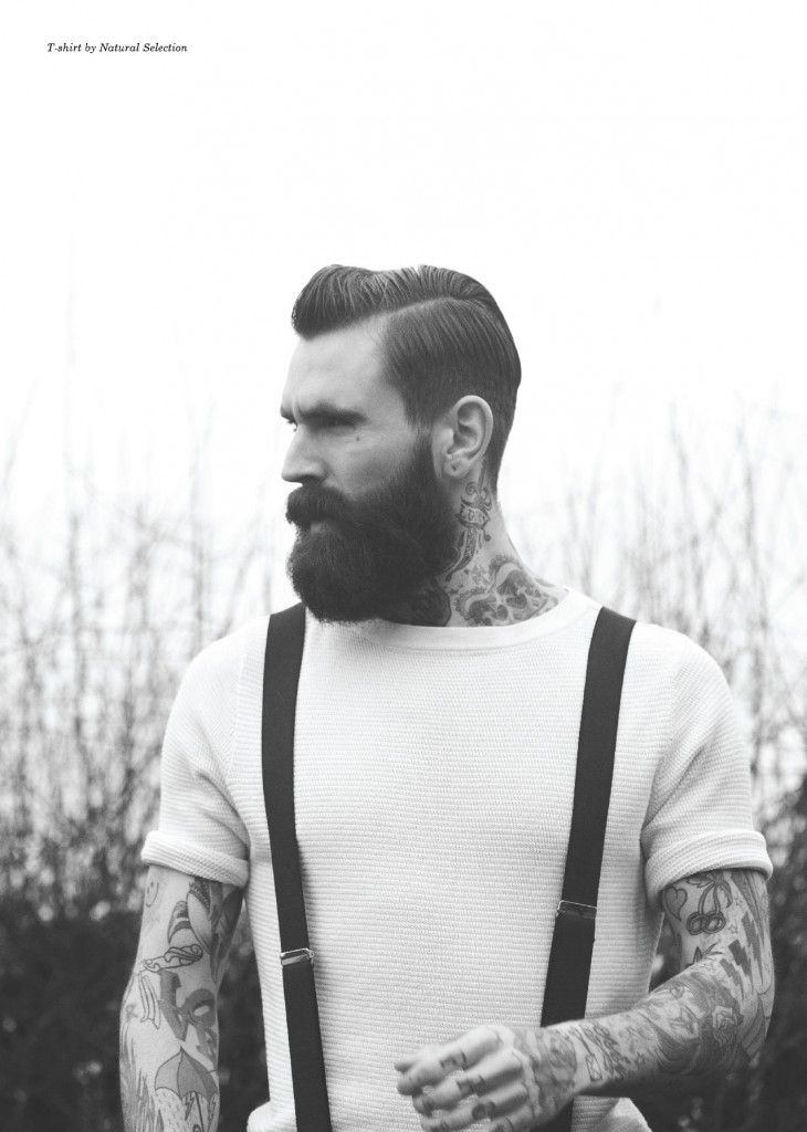 a close up portrait of a tattooed man with a beard. he is wearing a white tshirt and suspenders. he is looking at the camera with a serious expression. the photo is in black and white.