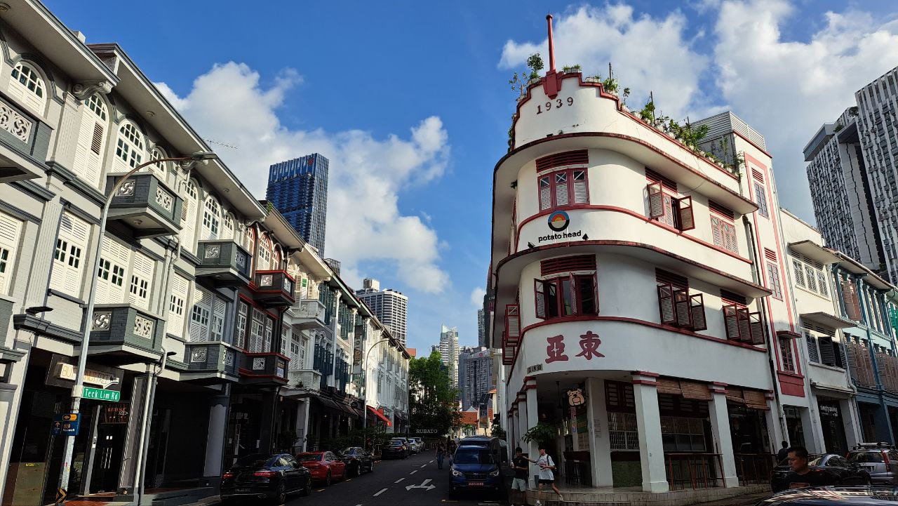 the image shows a vibrant street scene in singapore. the focus is on a row of beautifully preserved shophouses, characteristic of the city's colonial architecture. these traditional buildings, with their pastel colors and ornate details, are juxtaposed with modern highrise buildings in the background, showcasing singapore's blend of old and new.  the street is relatively quiet, with a few cars parked and people strolling along. one shophouse, prominently featured, is identified as "potato head," suggesting a restaurant or hotel. the overall atmosphere is peaceful and sunny, with a clear blue sky visible above. the image also includes street signs, indicating the location.