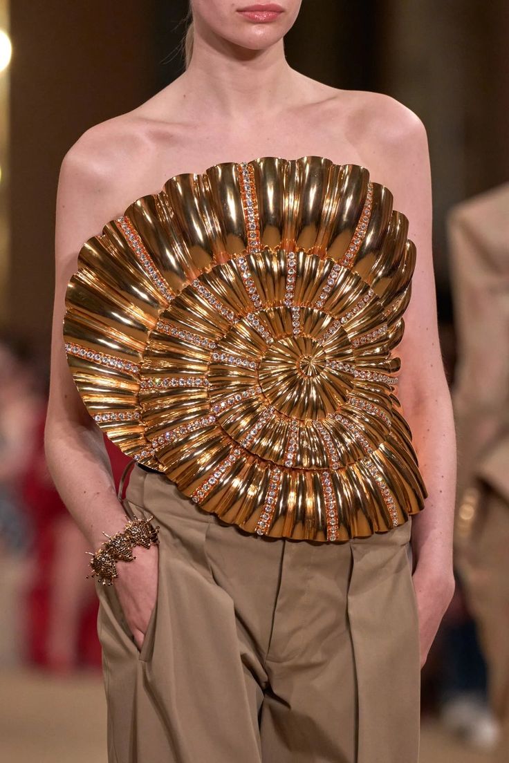 a model walks the runway in a gold jeweled top with a shell design. the top is a strapless piece and covers the model's chest and stomach. she is wearing brown slacks and has a large, jeweled bracelet on her wrist.