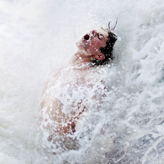 a young man is captured in a moment of pure exhilaration as he's engulfed by a powerful wave. the water explodes around him in a chaotic yet beautiful display of nature's force. his head is thrown back, mouth open in either a shout or a gasp, as he surrenders to the overwhelming sensation. the bright, natural light emphasizes the energy of the scene and the refreshing coolness of the water.