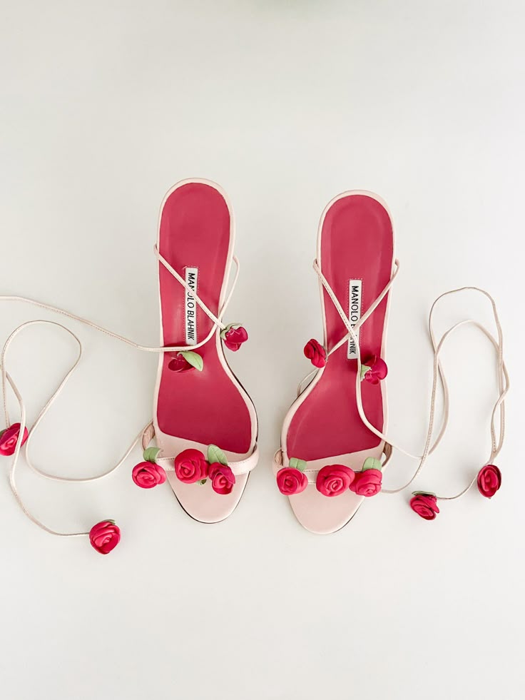 the image shows a pair of pink manolo blahnik high heel sandals with red rose accents. the sandals have a pink sole and straps that wrap around the ankle, adorned with small red roses. they are placed on a white background.
