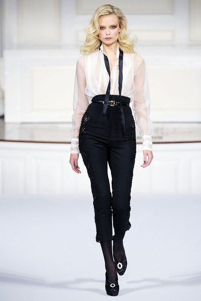 a female model walks down the runway in a white blouse, black pants, and black shoes. she is wearing a black belt with a gold buckle. her hair is styled in a sleek, straight look. she is walking confidently and posing for the camera.