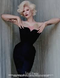 a glamorous black and white portrait of a blonde woman, possibly a model, posing in a black dress.  the image has a vintage or retro feel, with the woman's pose and styling suggesting a 1940s or 1950s aesthetic.  she has a sophisticated and elegant look. the background is a textured gray wall.  the woman has visible tattoos on her arms.  the image is styled to convey a sense of beauty and glamour.