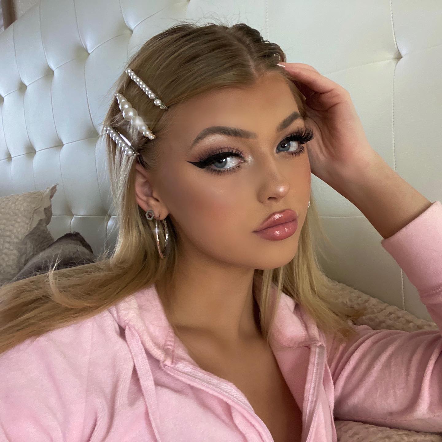 a blonde woman with blue eyes and glossy lips poses for a photo. she is wearing a light pink hoodie, hoop earrings, and several hair clips with pearls. she has her hand on her head and is sitting on a bed with a white headboard.
