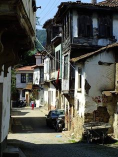 a narrow street in an old town, lined with traditional turkish houses. the houses are mostly white with wooden balconies and windows. some of the buildings are in need of repair. there are a few people and vehicles on the street. the street is lit by bright sunlight.
