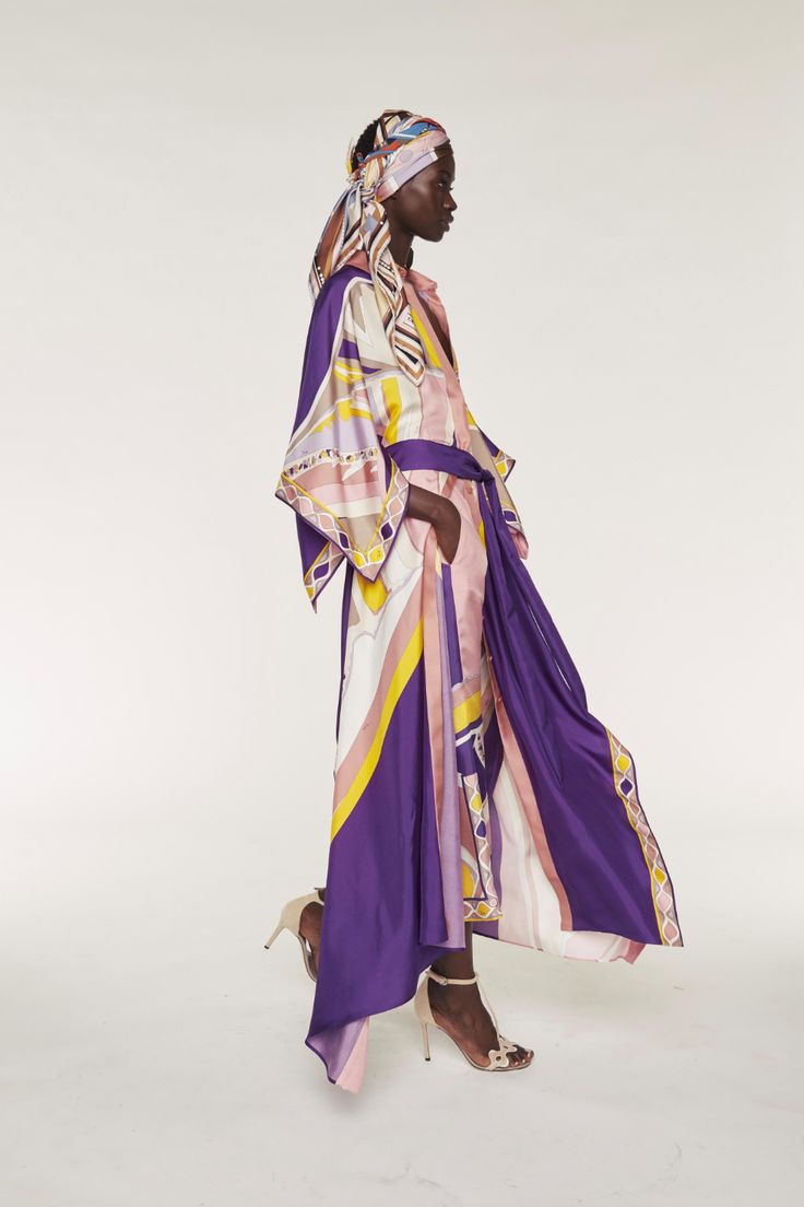 a full shot of a darkskinned female model showcasing a long, purple silk dress with a geometric print. the dress is belted at the waist and has long, wide sleeves. the model is also wearing a matching patterned headscarf and beige highheeled sandals. she is posing in a studio setting against a plain white background.  the overall aesthetic is elegant and sophisticated.
