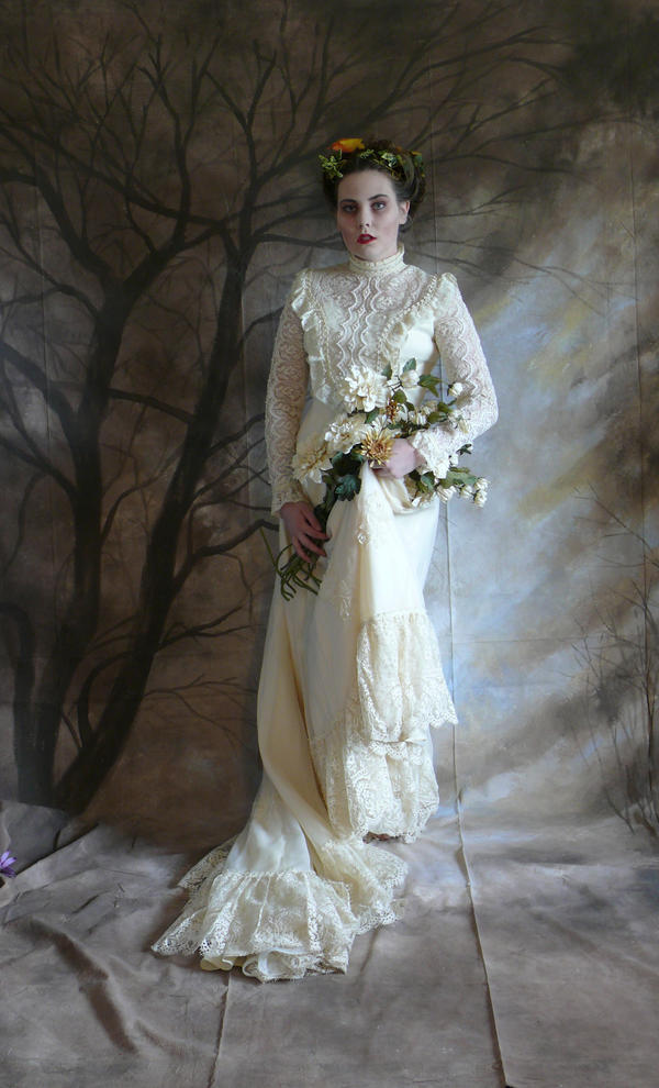a full shot of a woman in a vintage, offwhite lace wedding gown, posing against a dark backdrop of painted trees. she holds a bouquet of white and yellow flowers and is adorned with a floral crown. the overall mood is ethereal and somewhat somber.