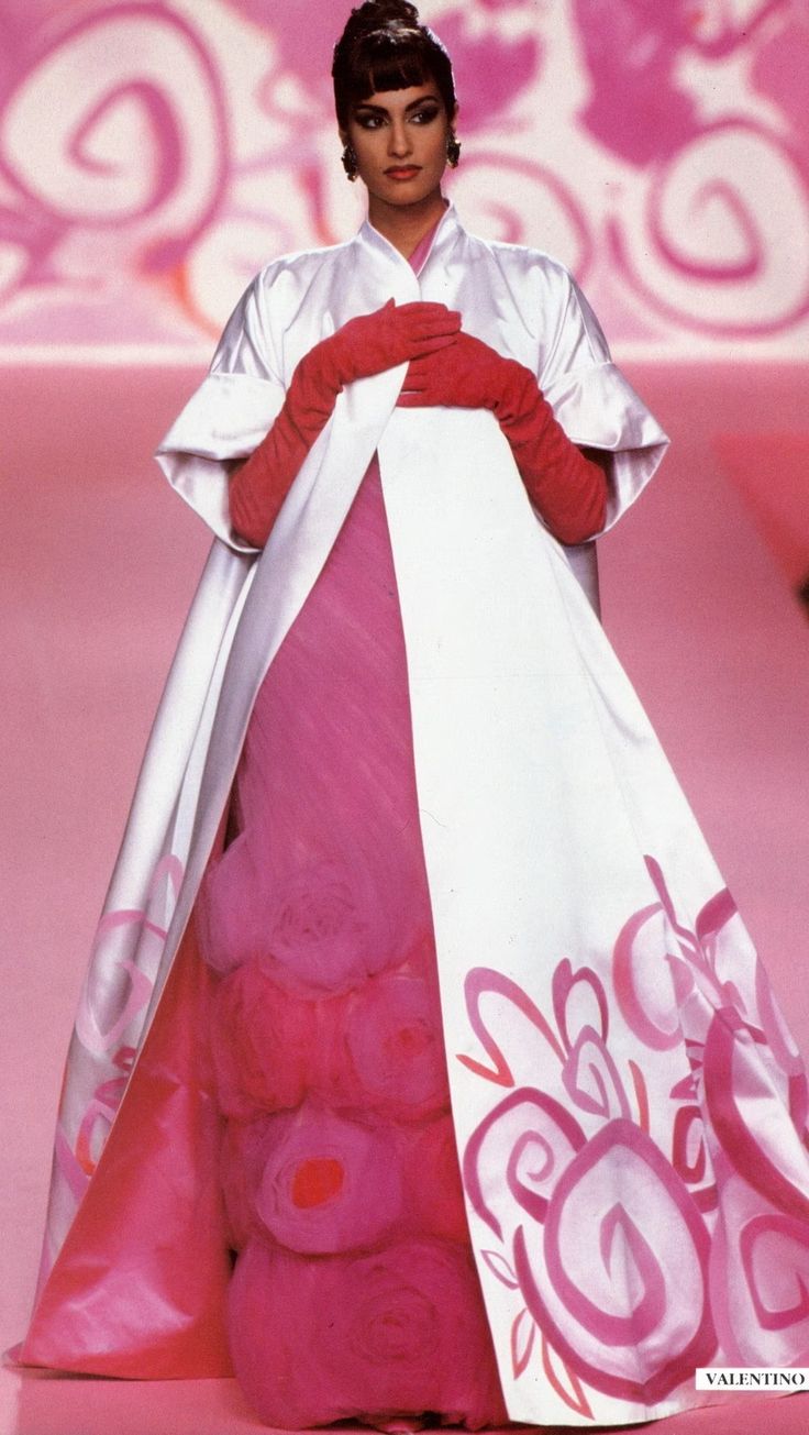 a model is seen on a runway during a fashion show wearing a pink gown with roses on the bottom underneath a long white jacket with a floral pattern at the bottom. the model is also wearing long pink opera gloves. the fashion show background is also pink and white. at the bottom right of the photo, the word "valentino" is visible.