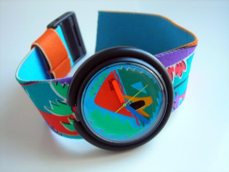 a closeup shot of a wristwatch with a black case and a colorful patterned band is positioned on a white surface. the watch face features a vibrant abstract design with orange, green, and blue colors. the watch band has a repeating pattern with elements of teal, purple, orange and green.