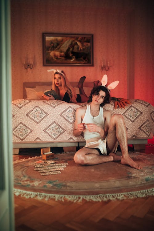 the image shows a young couple in a dimly lit, vintagestyle bedroom. the woman is lying on a bed with bunny ears on, partially hidden behind a patterned comforter. she appears to be reading a book. the man, also wearing bunny ears, is sitting on a round rug in front of the bed, holding a few playing cards.  there's a small pile of what looks like money scattered on the rug near him. the overall atmosphere is suggestive and dark, with a retro aesthetic, enhanced by the red lighting and vintage furniture including an antiquestyle bed and a painting on the wall. the scene feels both playful and slightly mysterious.