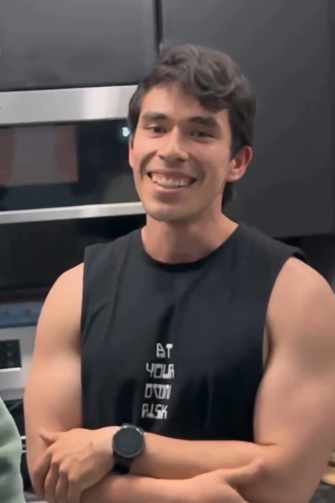 a man is smiling at the camera in a selfie. he is wearing a black tank top and a watch. his arms are crossed. the background is a kitchen.