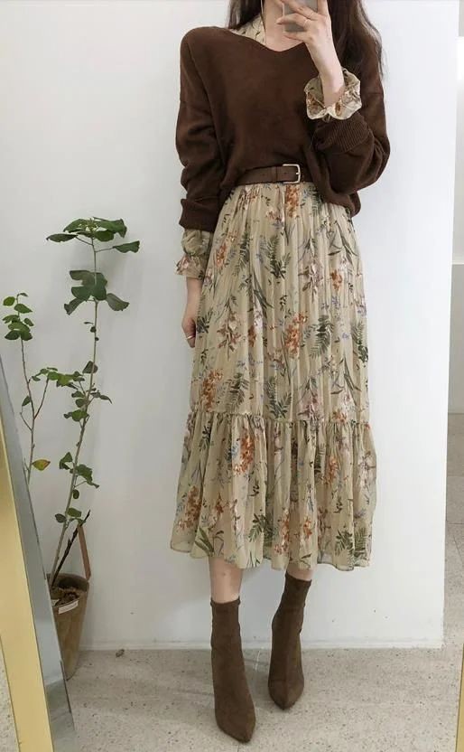 a woman is standing in front of a white wall, wearing a brown sweater over a floral dress with a brown belt, and brown ankle boots. there is a plant next to her on the left, and a mirror in the lower left corner.