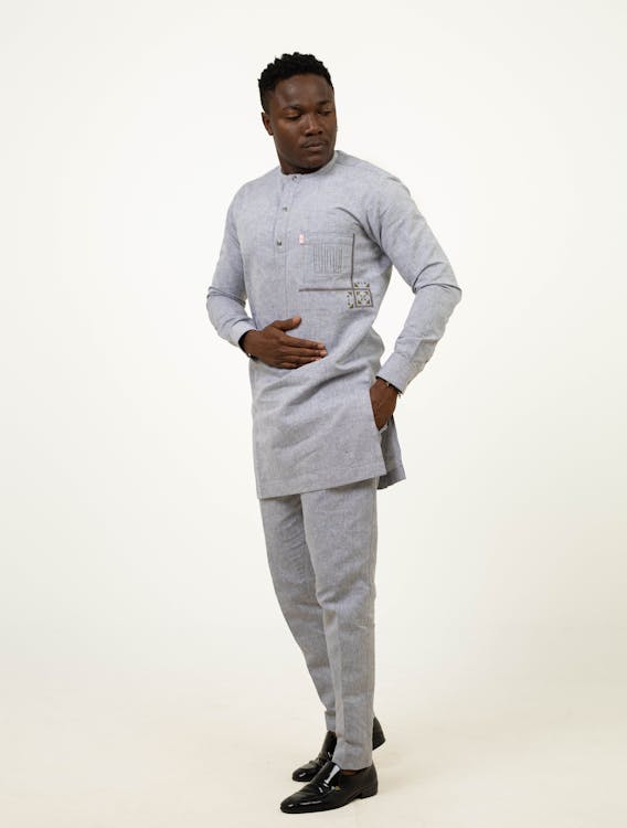 a man stands indoors wearing a light grey longsleeved shirt with a unique pocket design and matching pants. he also wears black shoes and looks down with one hand on his stomach.