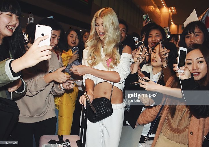 a blonde celebrity in a white outfit and pink top is surrounded by fans at an airport, who are holding up their phones to take pictures and get autographs.