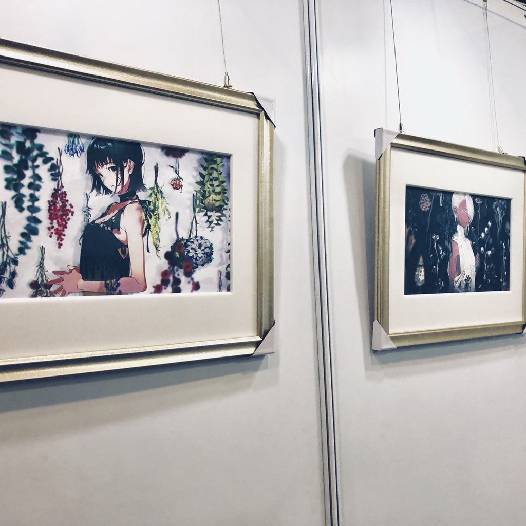 the image shows a section of an art exhibition. two framed artworks are hanging on a white wall. the artwork on the left features a girl with short black hair and a black top, standing amidst hanging flowers and plants. the artwork on the right is partially visible and shows a figure in a dark, ethereal setting with white flowers. the frames are goldcolored, and the lighting in the gallery is bright and artificial.