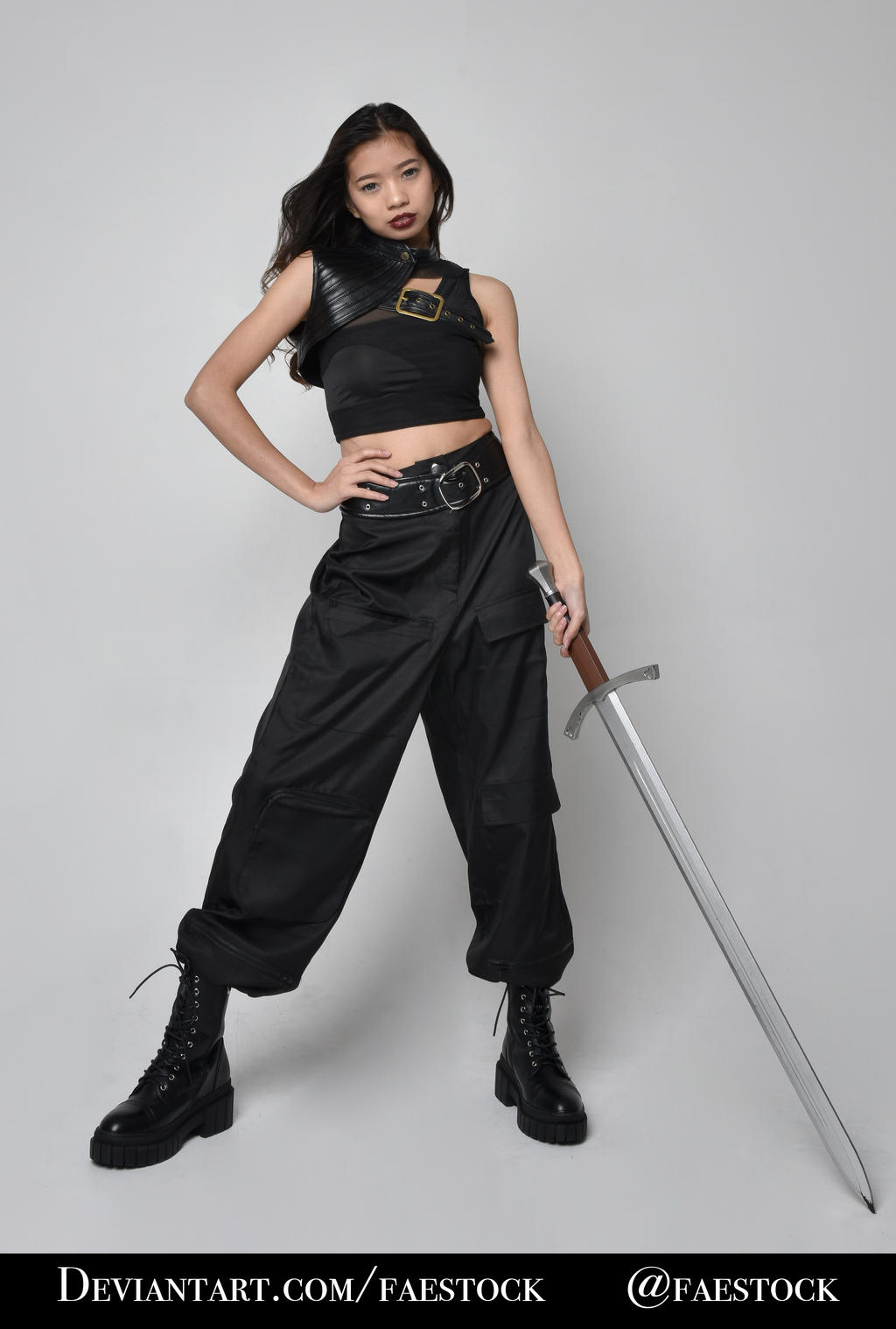 a fullbody studio shot of a young asian woman dressed in a fantasy warrior costume. she's wearing black cargo pants, black combat boots, a black sleeveless top, and a black leather shoulder armor piece with a gold buckle.  she holds a long silver sword, and her expression is serious and confident. the background is plain gray. the image is professionally lit and styled.  the photographer's watermark is visible.