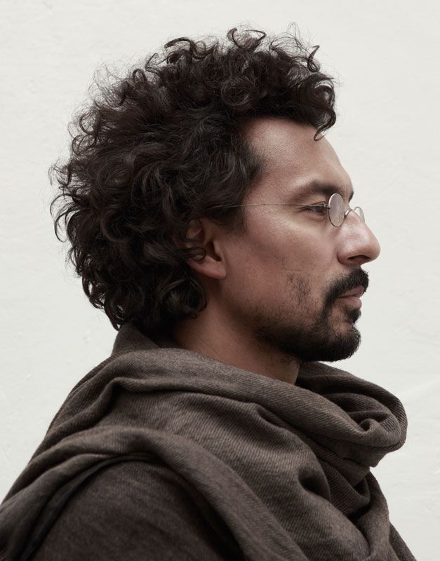 a side profile shot of a man with dark curly hair and a beard, wearing small round glasses and a brown scarf.