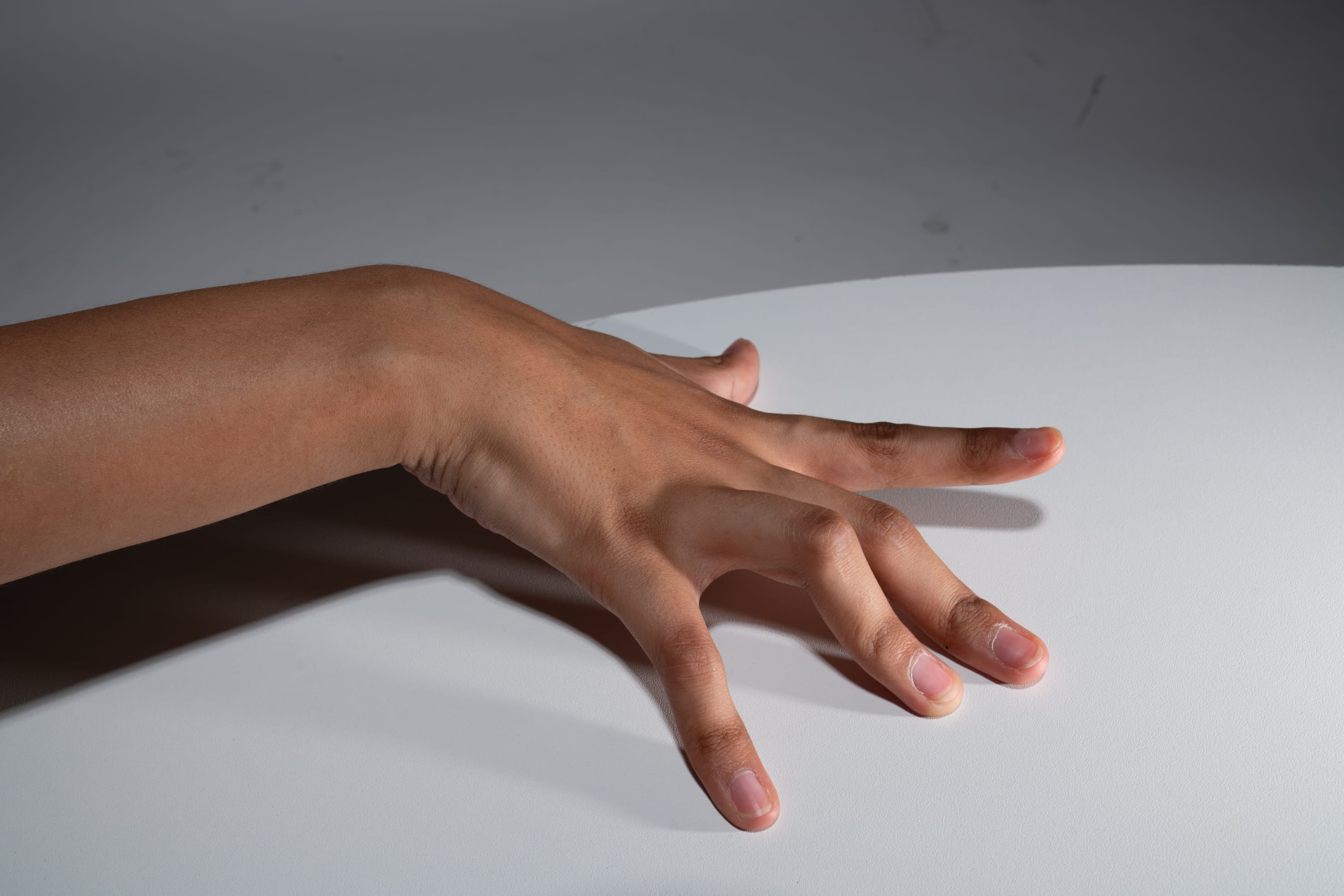 a hand with its fingers spread out on a white table. the hand is lit from the left side, creating a shadow on the right side.