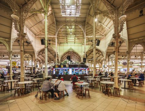 the image showcases the interior of lau pa sat (telok ayer market) in singapore.  the photograph captures the market's striking victorian architecture, featuring ornate castiron columns and a high, airy ceiling. numerous small tables and chairs are arranged throughout the central space, where many people are seated, enjoying meals at various food stalls. the bustling atmosphere and the intricate details of the building's structure are clearly visible, creating a vivid representation of this popular singaporean food market.  the lighting is bright, emphasizing the architectural features and the busy scene of people dining.