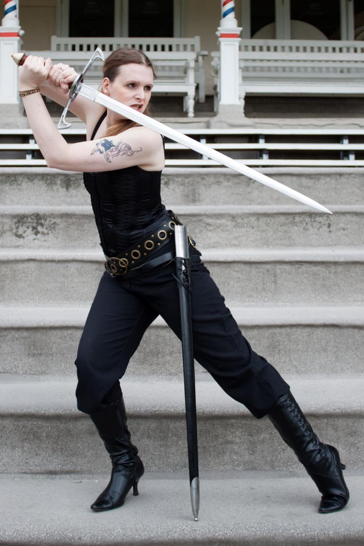a woman wearing a black corset and pants and boots poses with a sword. the woman is holding the sword in front of her as if she were going to strike.  she is standing on stairs outside.