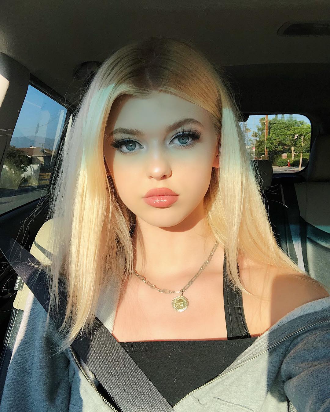 a selfie of a young woman with blonde hair and blue eyes, sitting in a car. she's wearing a gray sweatshirt and a black top, with a seatbelt across her chest. she has makeup on and is wearing a necklace. the car is parked, and you can see trees and a building through the window.