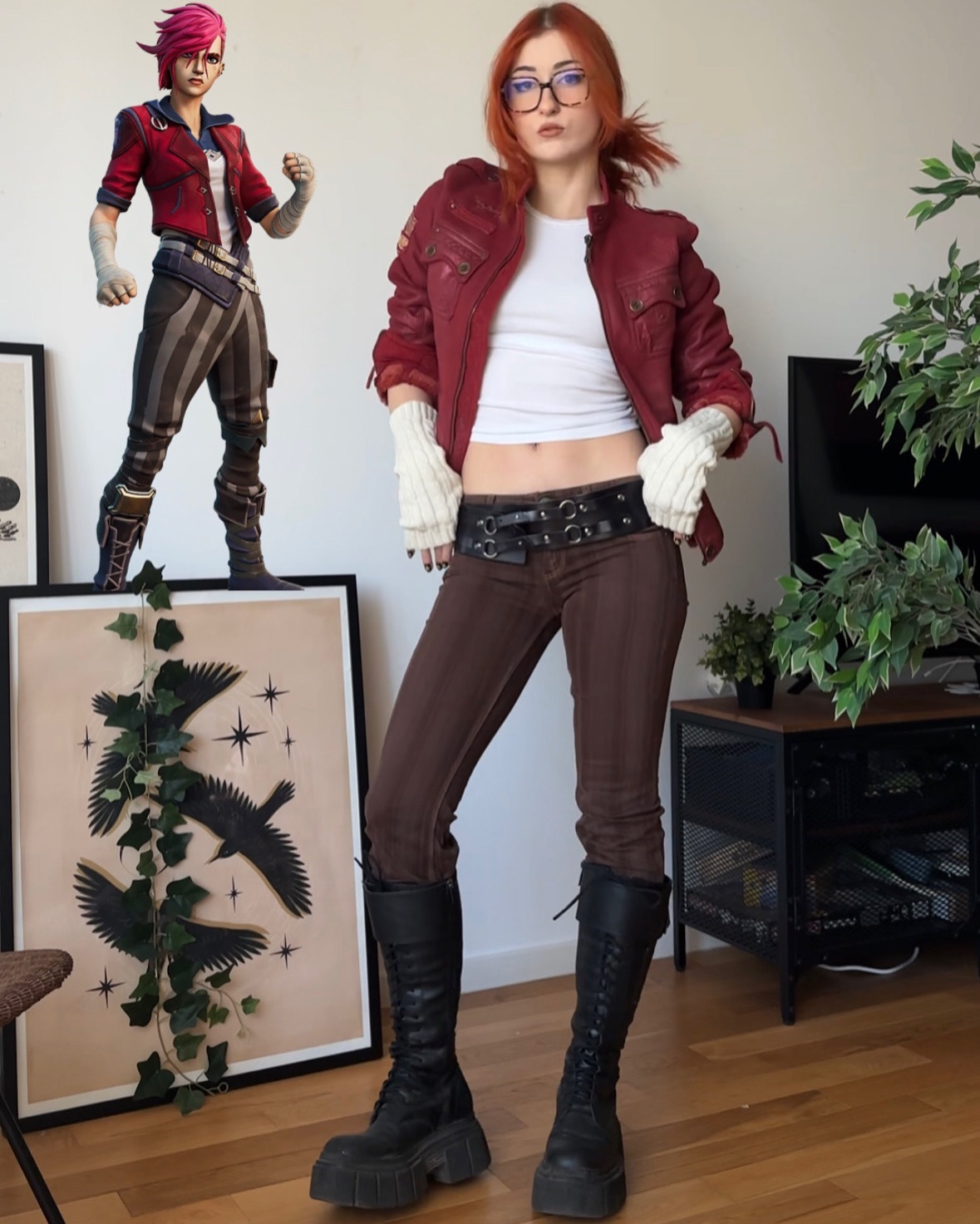 a woman is cosplaying as jinx from the video game riot games' arcane. she is wearing a red jacket, brown pants, and black boots. she has red hair and is wearing glasses. she is standing in a room with a picture on the wall and plants.