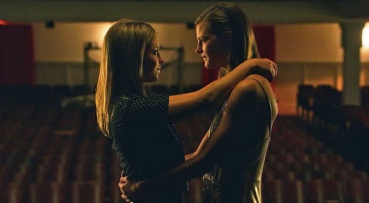 the image is a still from a film or television show, showcasing two young women embracing intimately in what appears to be an empty auditorium or theater. the lighting is dim and dramatic, focused primarily on the couple. they are closely intertwined, showcasing a tender and affectionate moment between them. the background is blurry but indicates a seating area for an audience.  the overall mood is romantic and emotionally charged.  the image strongly suggests a lesbian relationship.