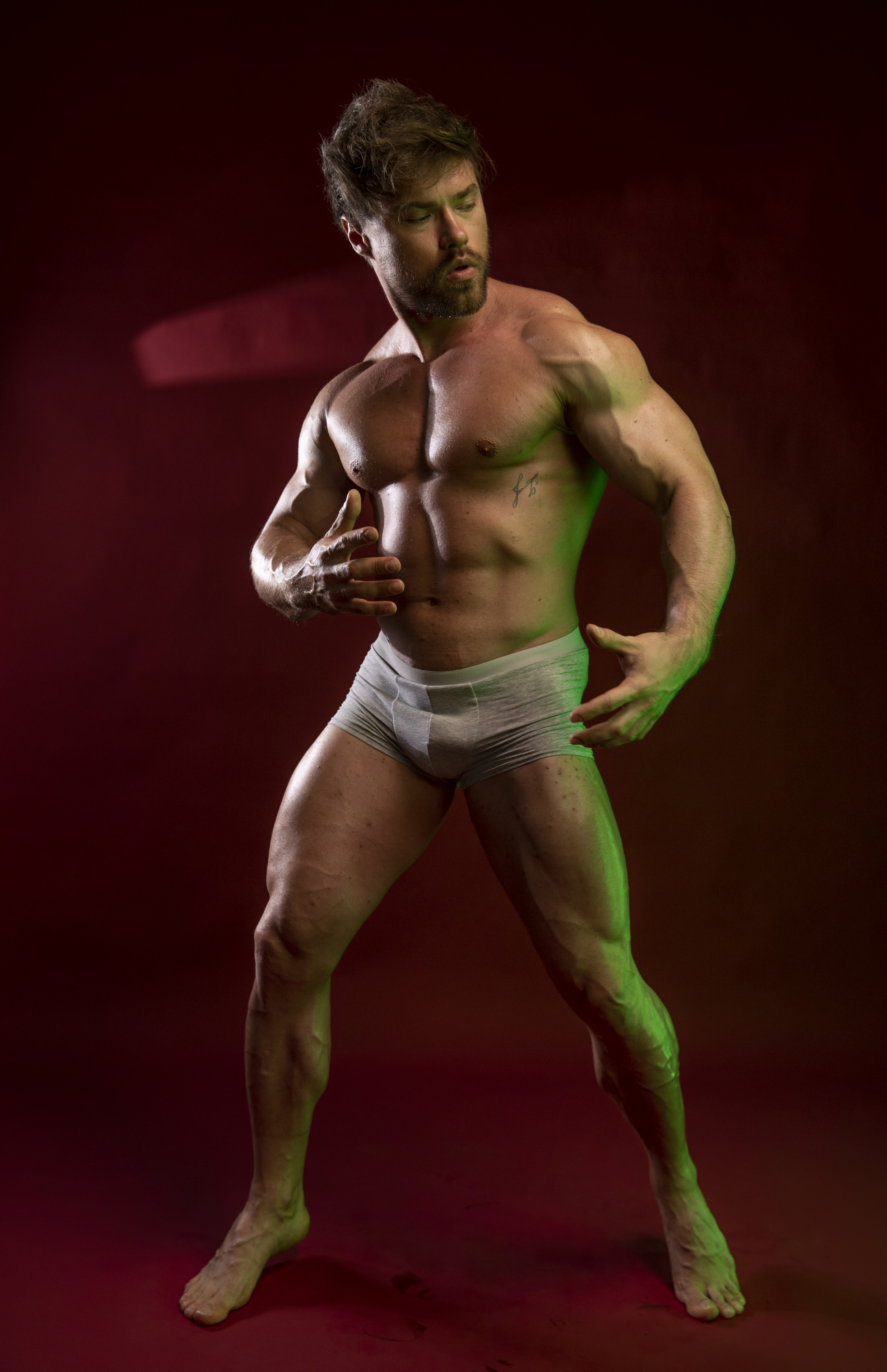 a fulllength studio shot features a shirtless, muscular man in underwear posing against a red background with green lighting. he has facial hair and a toned physique, showcasing his athletic and healthy appearance. he exudes confidence and sex appeal, positioned in a modern and artistic manner.