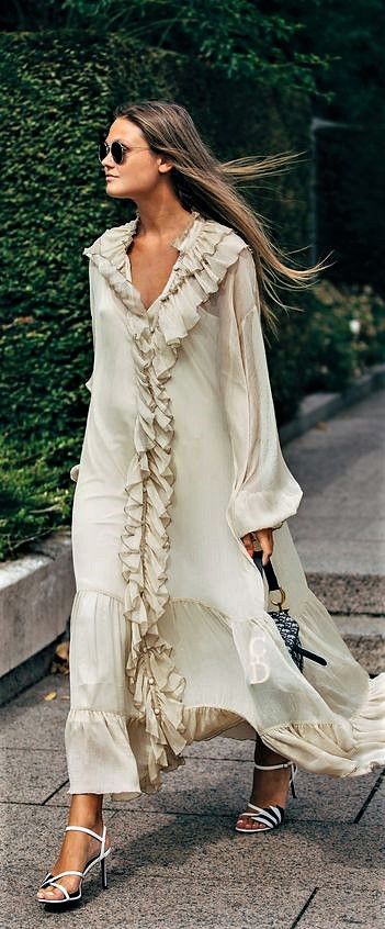 a fulllength shot of a stylish woman wearing a flowing, offwhite maxi dress with ruffles. the dress has long sleeves and a ruffled bodice.  she's accessorized with sunglasses, black and white heeled sandals, and a dior shoulder bag. the woman is walking on a sidewalk with lush green foliage in the background. her long hair is flowing in the wind. the overall style is elegant and summery.