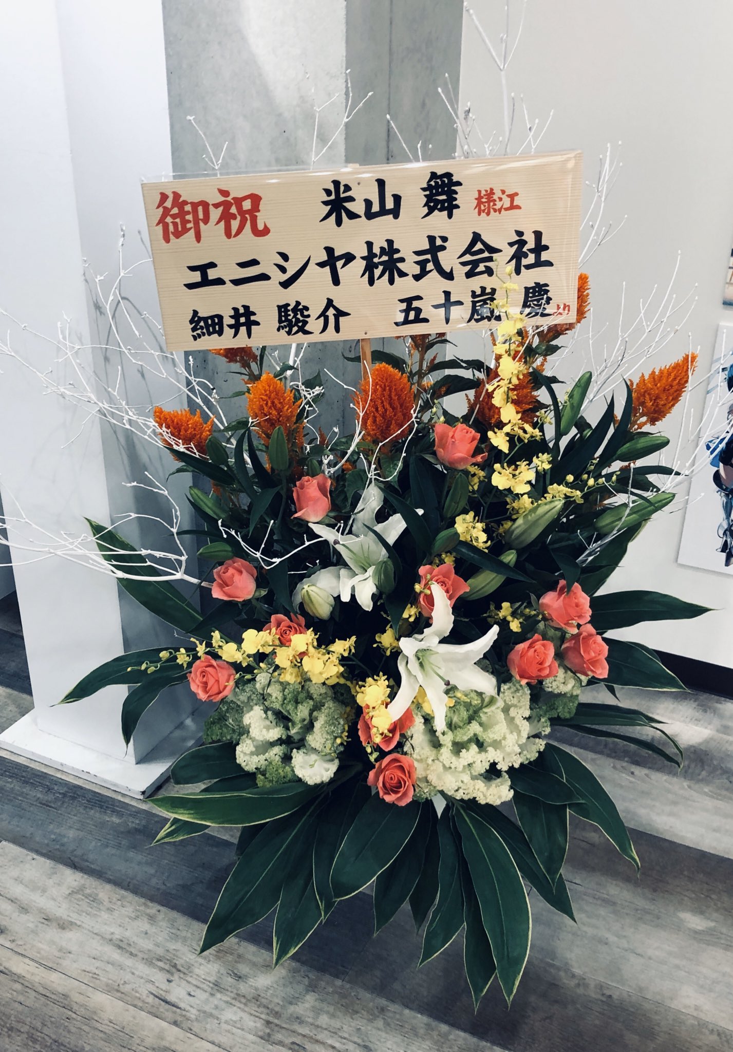 a vibrant floral arrangement stands in front of a white wall, accompanied by a congratulatory message in japanese. the flowers, predominantly pink, orange, yellow, and white, are arranged in a rectangular container. the handwritten sign, with black text on a beige background, celebrates the opening of a new business venture. the image suggests a joyous occasion and a thoughtful gesture of support.```