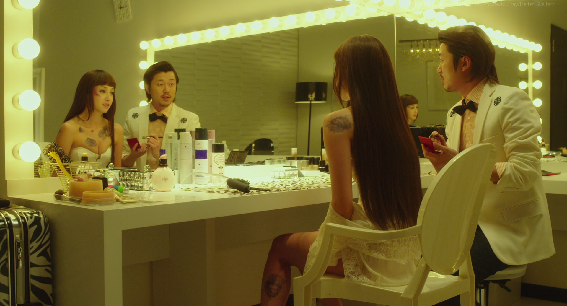 two couples are reflected in a large, brightly lit vanity mirror. the women are turned away from the camera, showcasing intricate tattoos on their backs and arms. they wear sheer white lingerie tops and dark stockings. the men are dressed in matching white tuxedo jackets and black bowties. one holds a compact while the other seems to be applying lip gloss. the vanity is cluttered with various beauty products, hair tools, and a