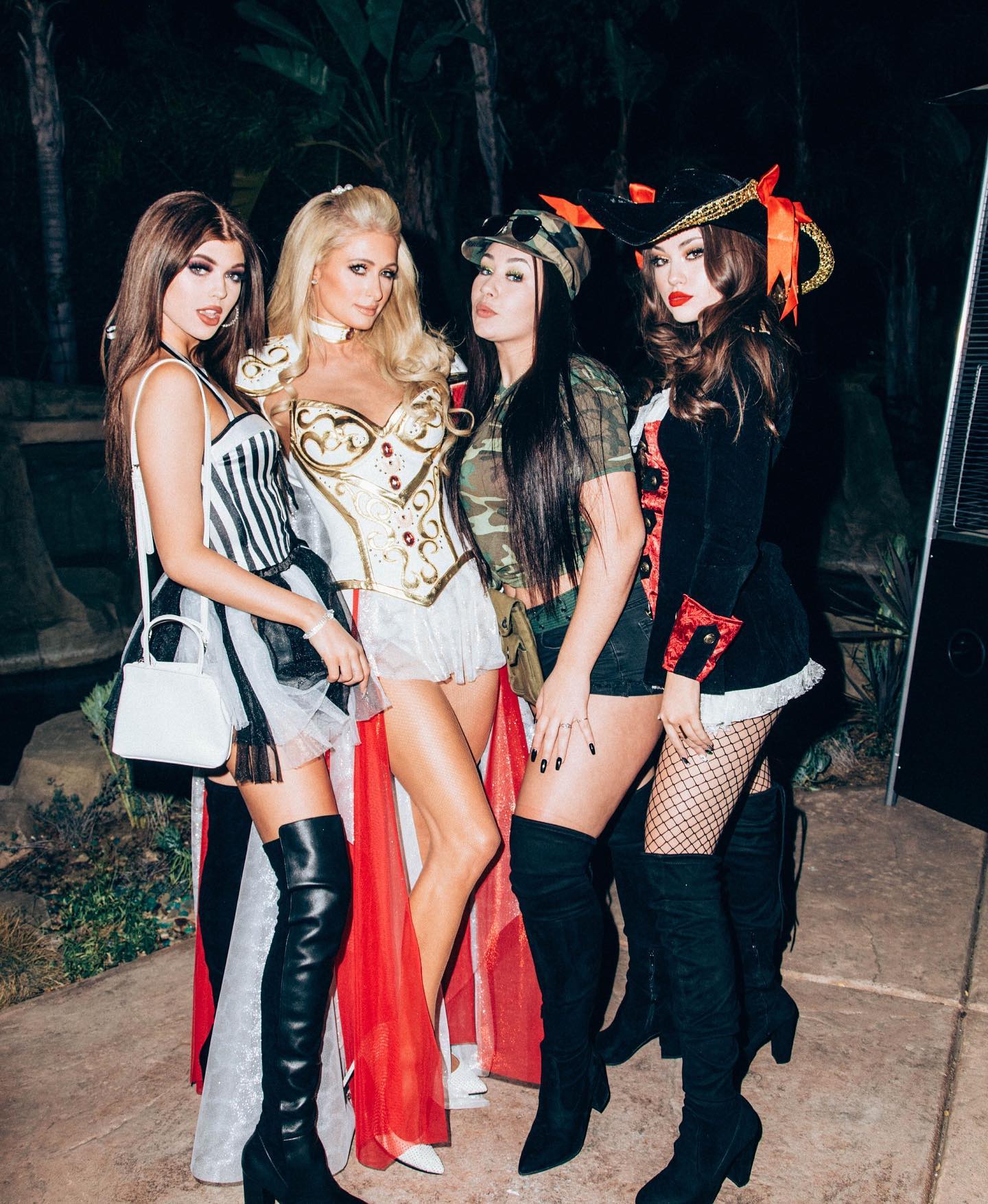 four women are posing in halloween costumes at an evening outdoor party. from left to right, the women are dressed as a referee with black and white striped top and black boots, a princess, a soldier in a camo hat and outfit with black boots, and a pirate with red lip stick wearing fishnet stockings and black hat with orange accents. they are standing together in a group with trees and foliage visible in the background.