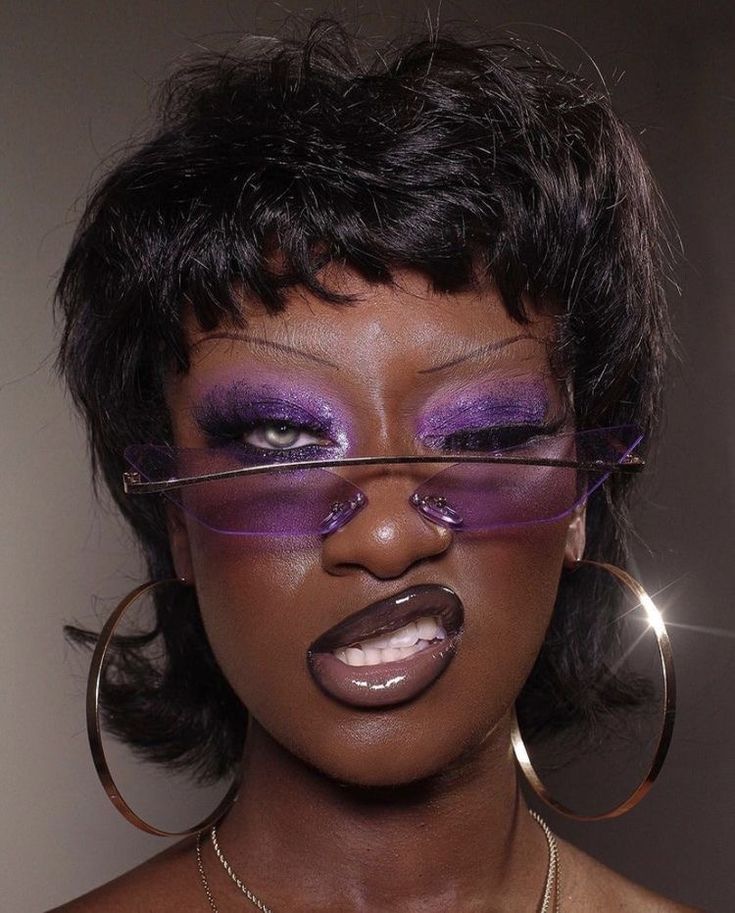 a close up portrait of a woman looking at the camera. she has short black hair, a purple eyeshadow, a glossy light purple lip, and wears sunglasses with a gold chain choker.