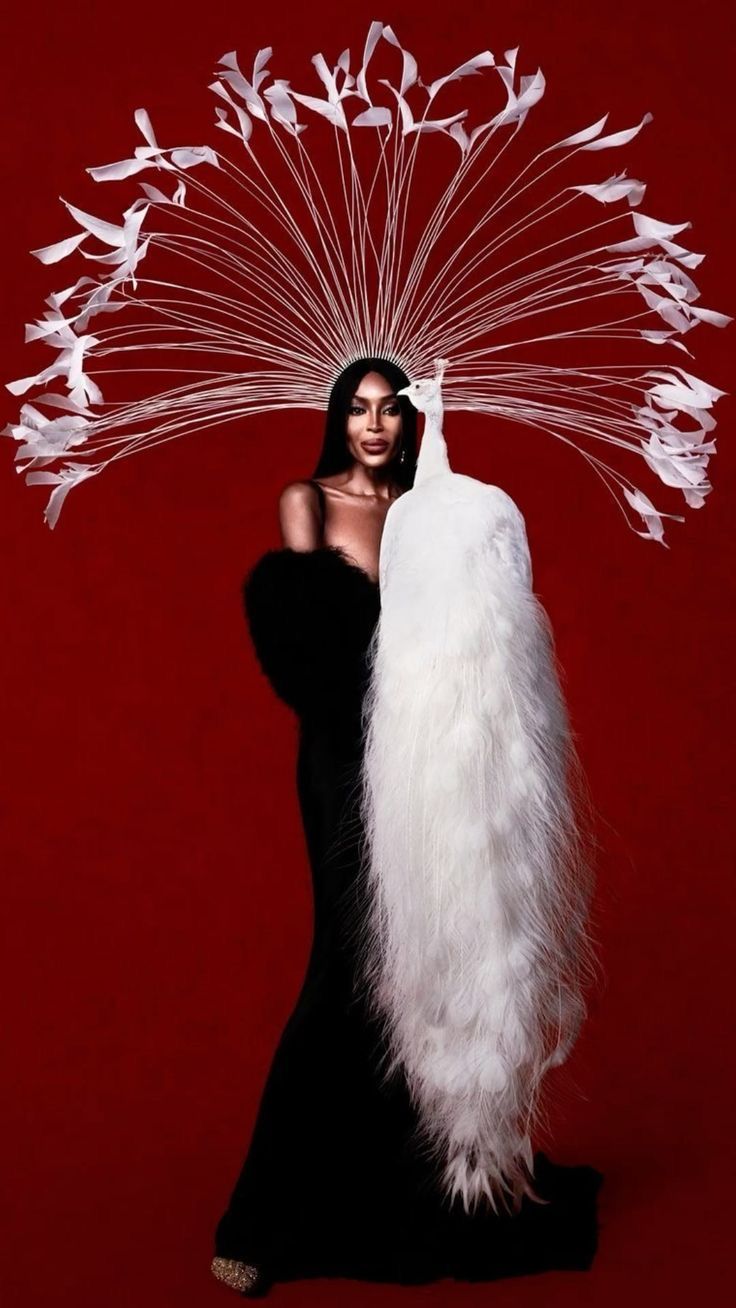 a woman in a black dress is standing against a red background. she has a white peacock draped over her shoulder and a large white feather headdress.