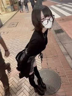 a woman in a black outfit and sunglasses walks along a city street with a mask over her mouth and nose.