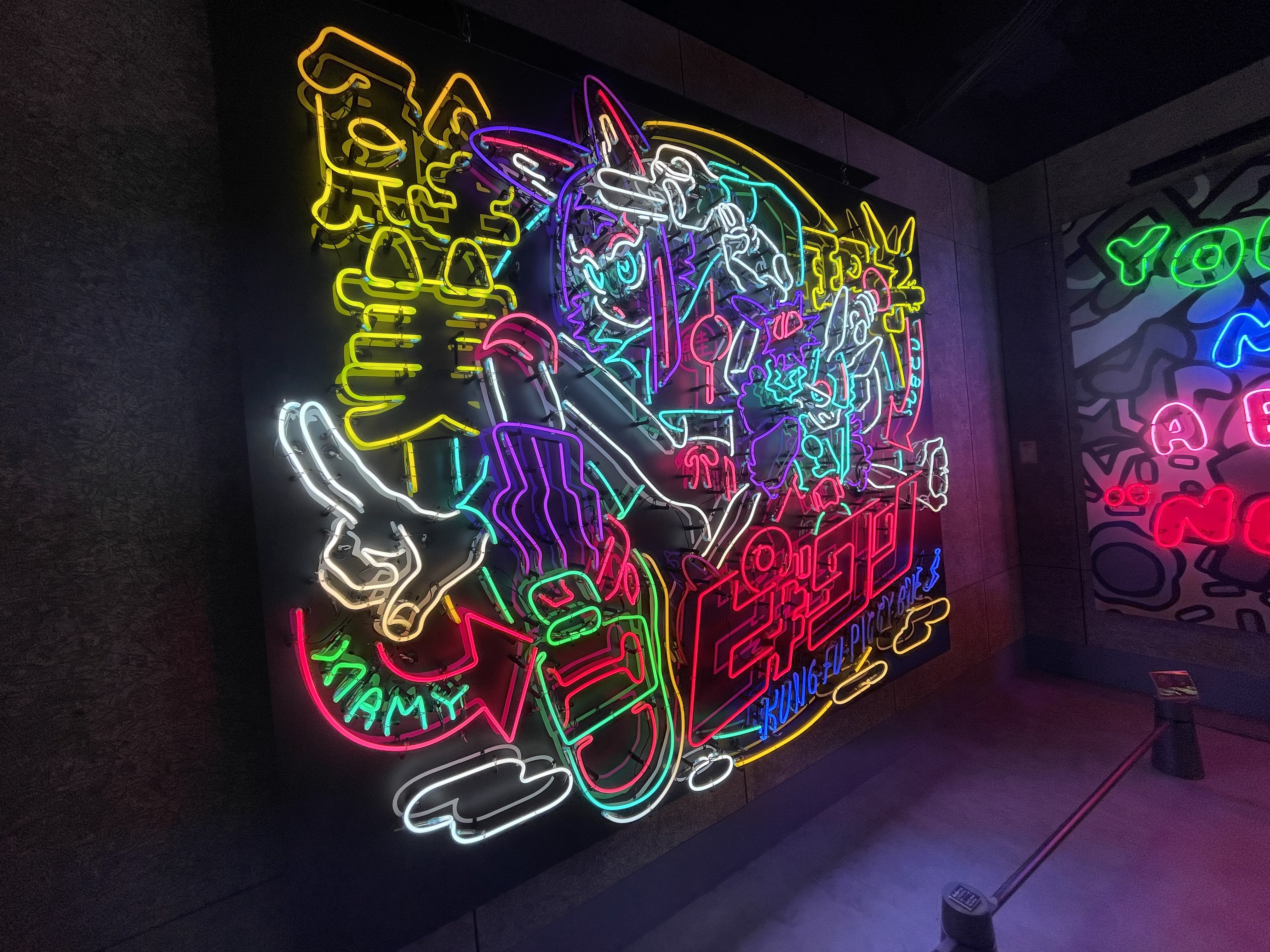 the image showcases vibrant neon artwork within an indoor setting, likely an art gallery or museum. the artwork features a dynamic composition with a central figure in a cartoonish, streetartinspired style. bold lines, bright colors, and japanese characters contribute to the energetic aesthetic. the neon lighting illuminates the artwork and casts a colorful glow on the surrounding space.  the perspective is from eye level, providing a direct view of the artwork and its impact.