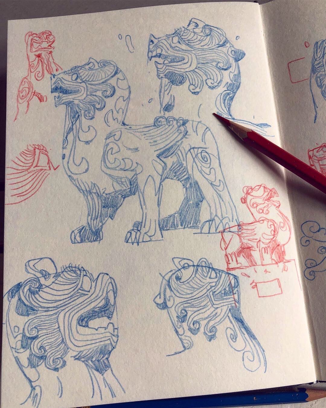 a sketchbook page featuring several sketches of a komainu, a mythical lionlike creature often found guarding temples in japanese culture. the sketches explore different angles and details of the creature, including its mane, teeth, and pose. the drawings are done in blue pencil with some red markings, and there is a red colored pencil resting on the page.