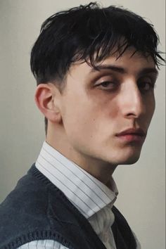 a young man with dark hair and sharp features gazes intensely at the viewer. his hair is styled in a modern cut with a wet look. he wears a blue vest over a white pinstriped shirt.  his expression is serious, contemplative, and slightly intimidating.  the lighting is soft and natural, highlighting his features without harsh shadows. the background is a neutral beige tone.