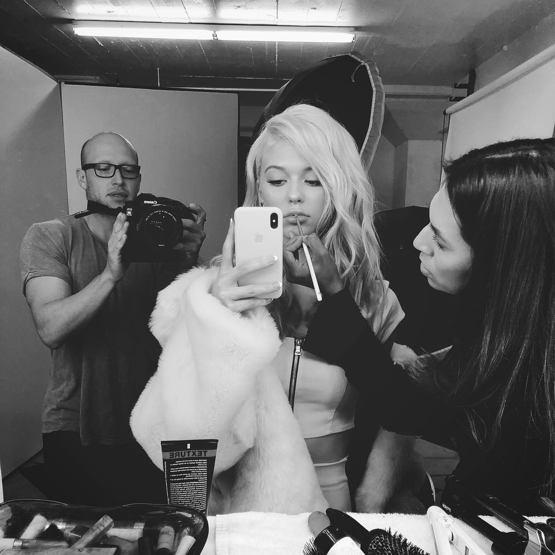 a black and white image shows a blonde woman in a makeup chair, getting her lip makeup applied by a makeup artist, while taking a selfie with her iphone. a photographer stands in the background with a camera. a white fur coat is draped around her, and a makeup kit is visible in the foreground, along with "texture" branded hair products.