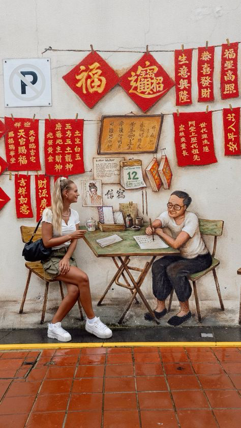 the image depicts a vibrant street mural in singapore. the mural features a traditional chinese storefront scene with red lanterns and calligraphy displaying auspicious phrases. a young woman in modern clothing sits at a table across from an elderly man writing at a table. the scene is richly detailed, showcasing a blend of traditional and modern elements, offering a glimpse into the cultural heritage of the area. the style of art is illustrative and detailed. the mural is painted on a white wall. the overall setting is a street with red tiled pavement visible at the bottom of the picture.