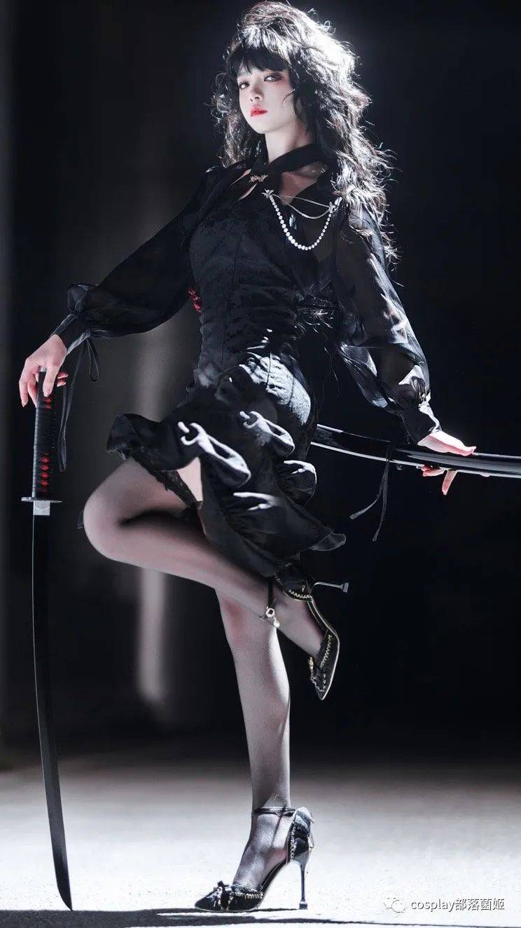 a woman in a black dress and high heels poses with two swords. she has black hair with bangs and wears a pearl necklace. she wears black stockings. one sword stands upright, the second one she holds horizontally in her hands.