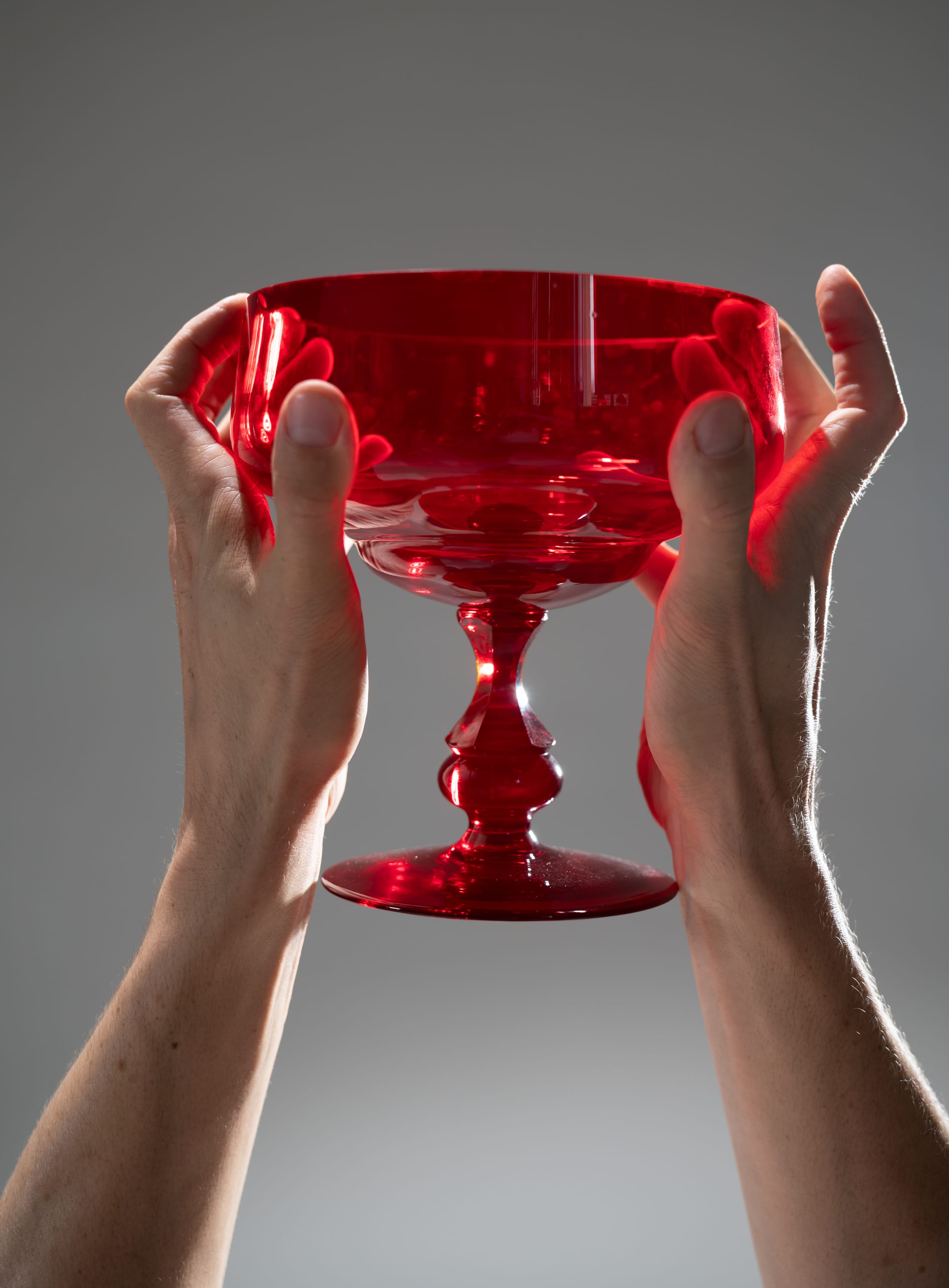 two hands are holding up a red glass goblet against a minimalist grey background. the goblet is illuminated by studio lighting.
