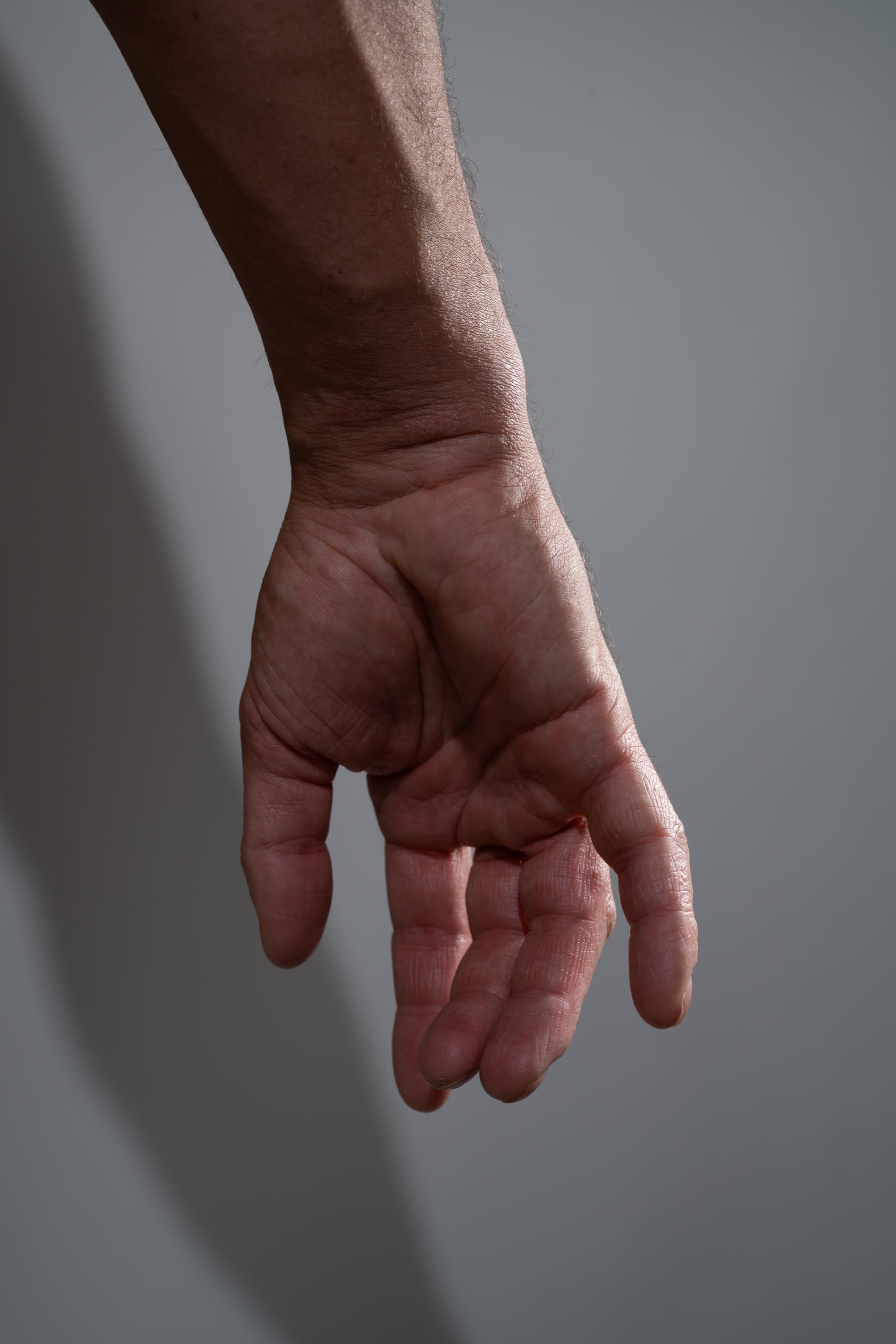 a closeup image of a man's arm and hand extended downwards against a light gray background. the hand is relaxed with the fingers slightly curled inwards. the lighting is soft and natural, creating subtle shadows on the wall. the image focuses on the details of the skin and the musculature of the arm.