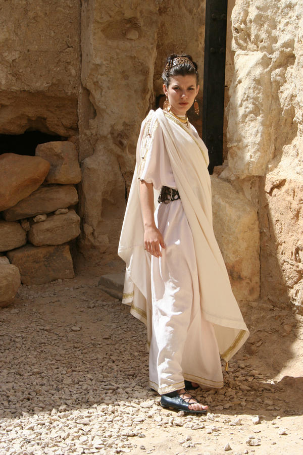 a fullbody shot of a woman dressed in a white stola, a traditional garment from ancient rome or greece. she is standing in front of a stone ruin. the woman's hair is styled in an updo. she is wearing sandals and jewelry. her expression is serious. the setting is outdoors and sunny. the image is evocative of a historical or costume context.