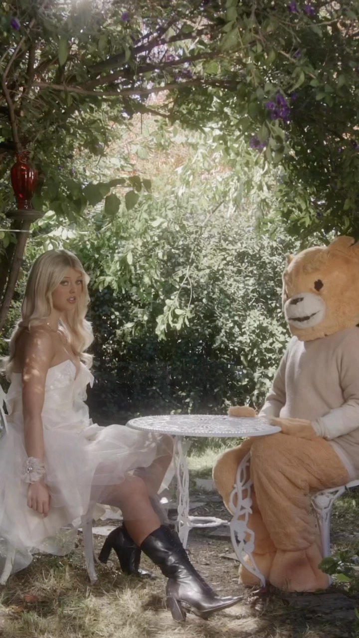 a woman in a white dress and black boots sits across a white table from a person in a bear costume in a garden setting.