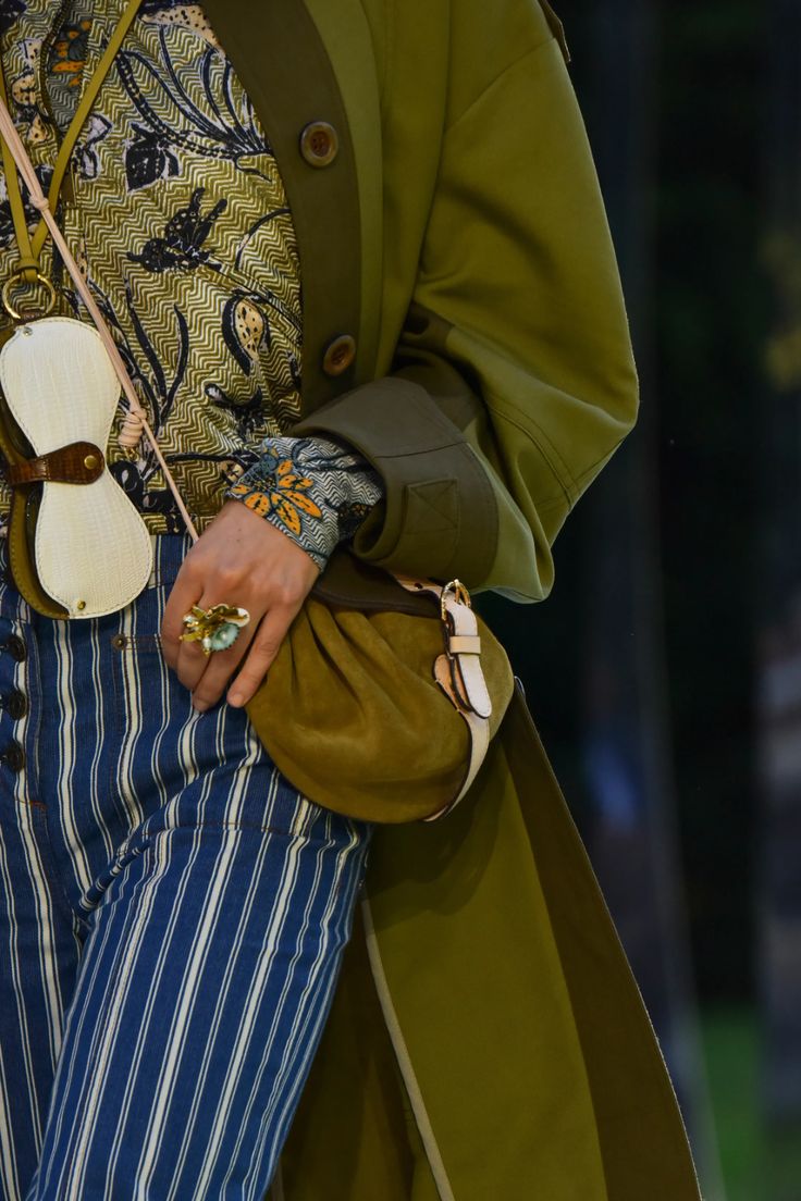loseup view of a fashion model showcasing a stylish ensemble. the model is wearing a long olivegreen coat, a patterned blouse with floral and paisley designs in shades of green and black, and blue and white pinstriped pants. a noticeable accessory is a green suede handbag with light beige straps. a unique ring is also visible on one of the model's fingers. the overall aesthetic is sophisticated and blends various patterns and textures, indicative of highfashion runway styling. the background is blurred, focusing attention on the clothing and accessories.