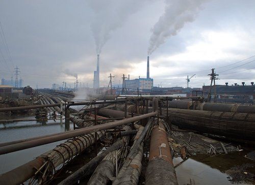 the image shows a scene of heavy industrial pollution. in the background, several smokestacks from factories belch out dark smoke into the overcast sky. in the foreground, numerous large, rusty pipes are strewn across a muddy area, many seemingly abandoned or derelict.  water is visible in the foreground, possibly polluted by the industrial runoff. the overall impression is one of environmental degradation and industrial decay. the scene appears to be in a postsoviet industrial area, likely in russia. the image is bleak and conveys a strong sense of environmental damage.