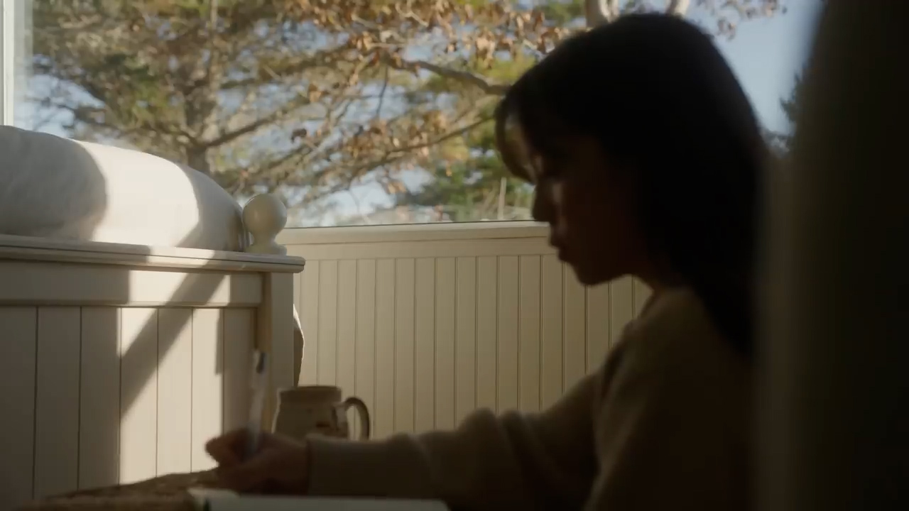 a woman sits in a sundrenched room in profile, writing in a notebook with a pen in hand. she is positioned beside a window offering a glimpse of trees and  a white bed is to her other side. the scene evokes a sense of peace and quiet contemplation.
