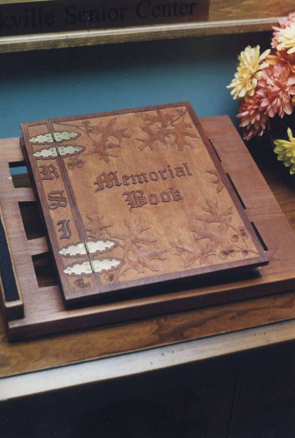 the image shows a wooden memorial book displayed on a wooden stand. the cover of the book is elaborately carved with a floral design and the words "memorial book" in gothic lettering. there are decorative brass accents securing the spine. the book is placed on a tiered wooden stand. in the background, there is a plaque with "oakville senior center" inscribed. a colorful bouquet of flowers sits to the right of the book stand. the scene is set on a table in what appears to be an interior setting.
