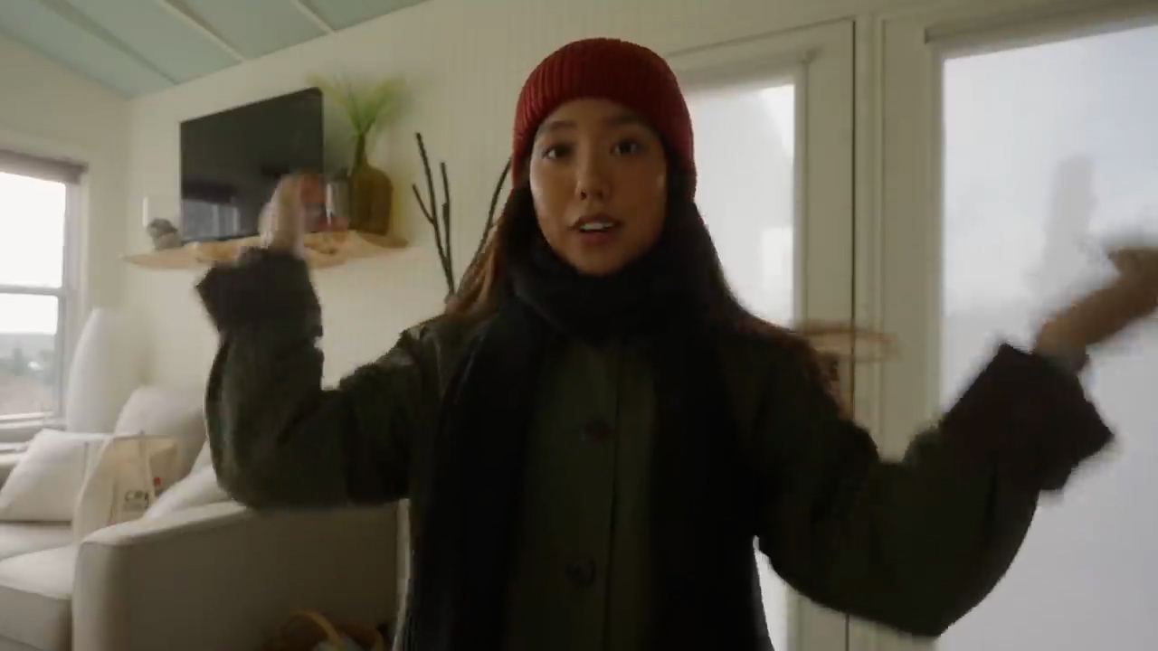 a young woman with long dark hair is standing in a living room. she is wearing a green jacket, black scarf, and a red beanie. she is talking and gesturing with her hands. the room is bright and airy with white walls and light wood floors. there is a couch, a tv, and a few other pieces of furniture in the room.